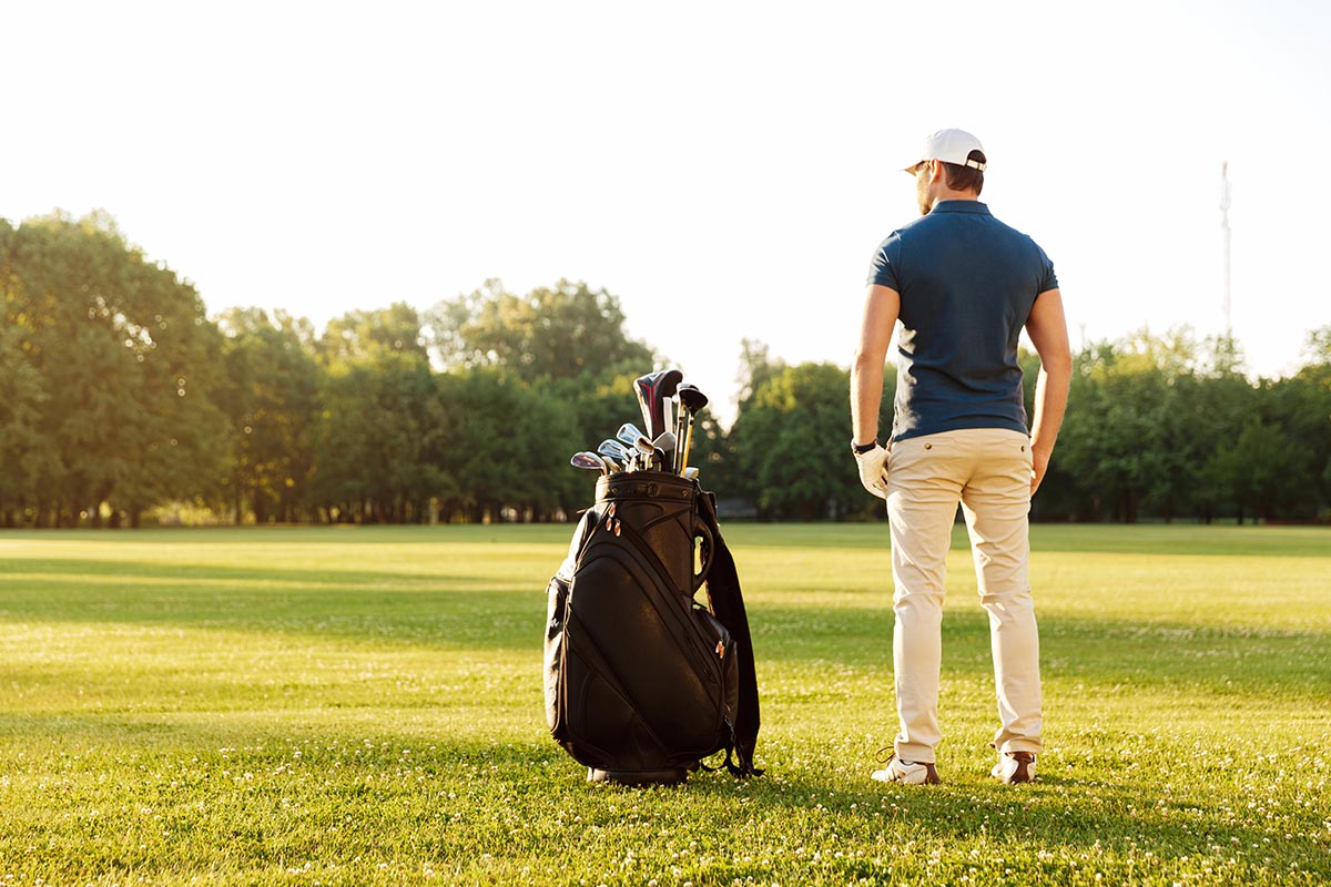 Back view of a young man standing on a green field with golf bag